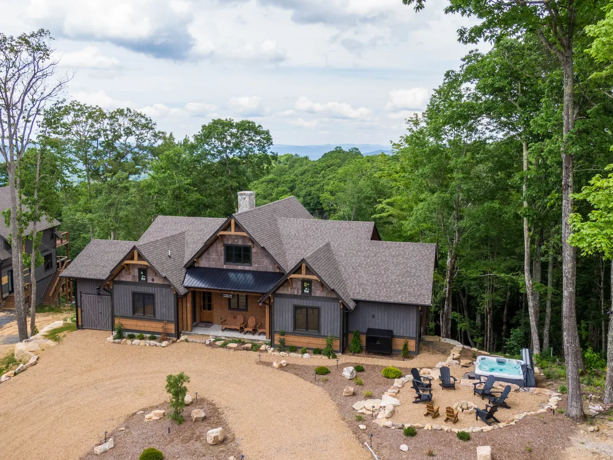 The image features the cabin's grand entrance with a welcoming covered porch, a bubbling hot tub perfectly positioned for scenic views, a cozy fire pit surrounded by inviting chairs, and beautiful natural stone landscaping nestled amidst the tranquil forest. The warm glow of the setting sun adds to the peaceful, mountain retreat ambiance.