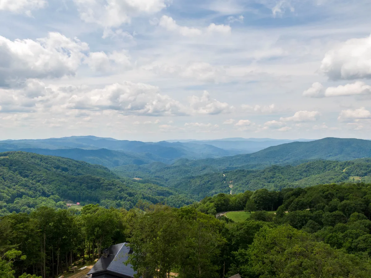 Expansive, lush green mountains and rolling hills stretch beneath a wide, cloudy blue sky, providing a breathtaking panoramic view typical of the High Country landscape.