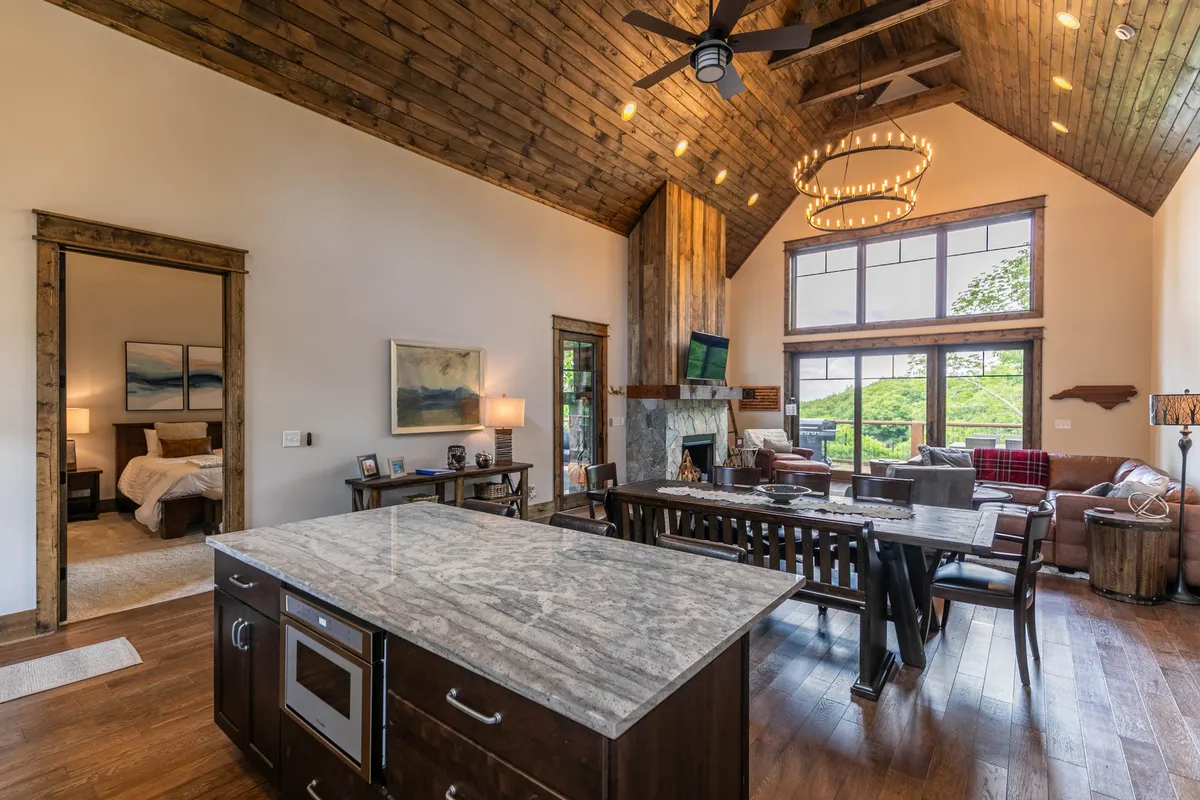 A wide-angle view encompassing the kitchen, dining table, and living area, all unified by a consistent, cozy design. The kitchen island, comfortable seating, and a stone fireplace accentuate the room’s social and functional versatility, while expansive windows bathe the entire space in natural light, further tying indoors to the mountain landscape.