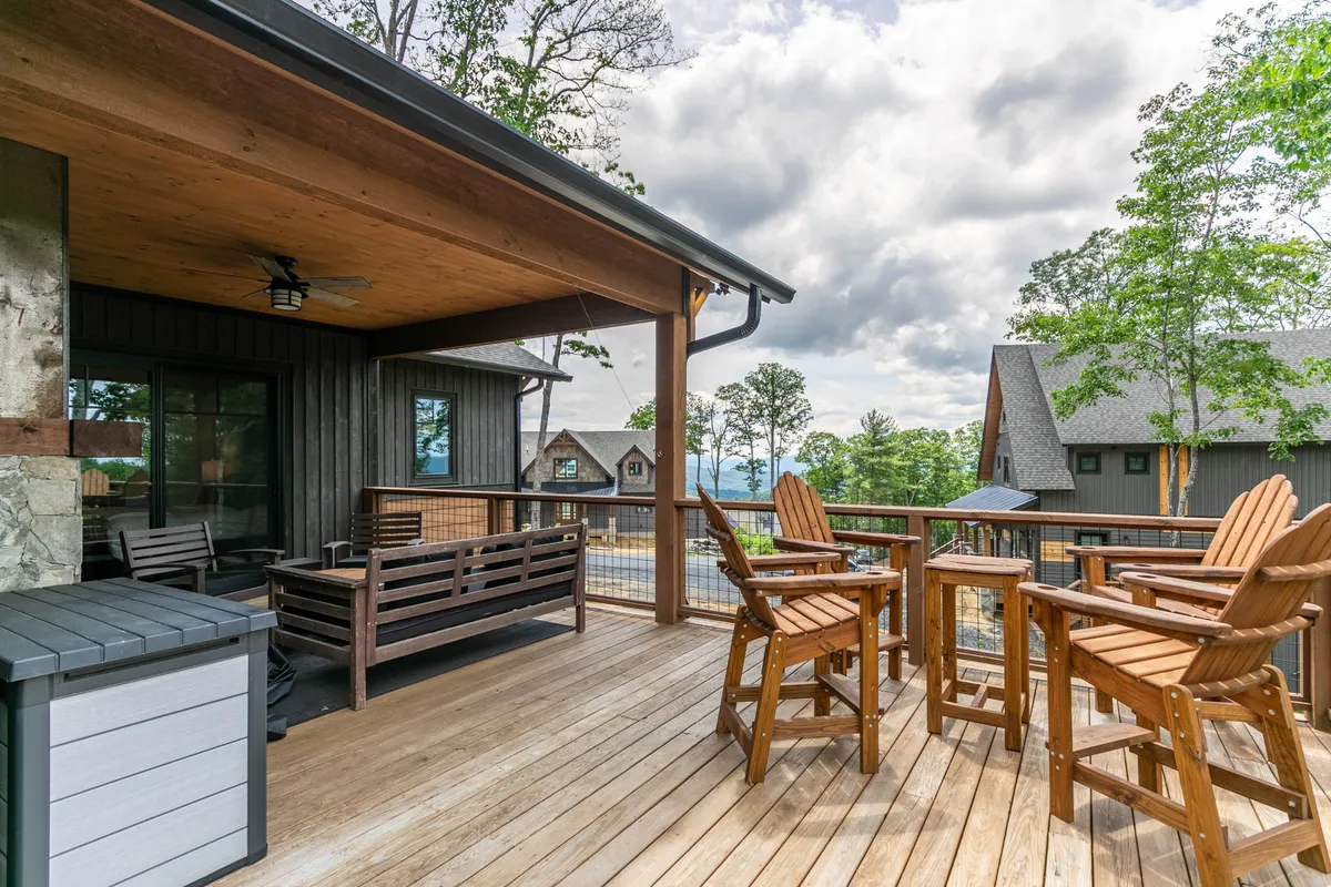 This image portrays a spacious wooden deck equipped with bar stools and comfortable outdoor seating, surrounded by lush trees and offering a distant view of rolling hills. The scene is tranquil, with the deck positioned perfectly for enjoying drinks, meals, or serene moments in nature.