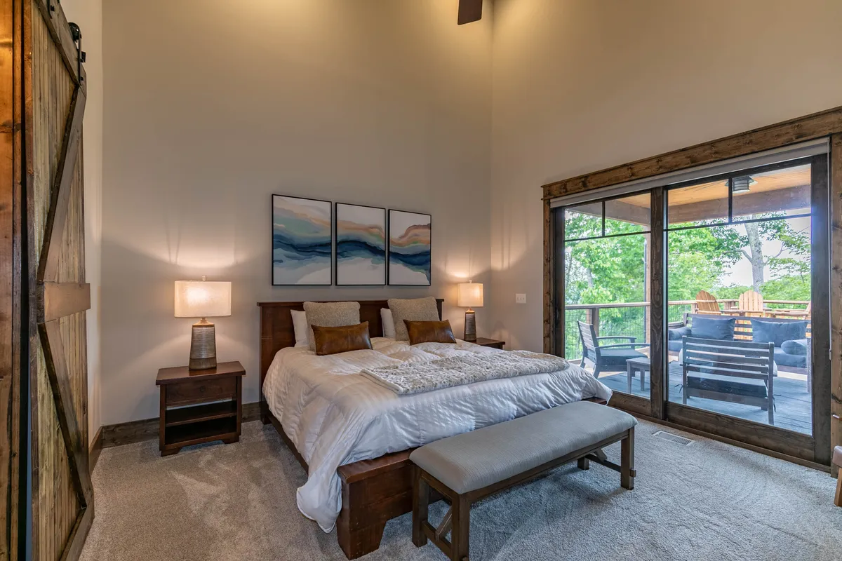 A serene bedroom featuring a large bed with a neutral-toned comforter, flanked by nightstands and lamps. Artwork above the headboard and a sliding glass door bring in natural light, while a bench at the foot of the bed adds comfort. The overall atmosphere is calm and inviting, perfect for restful nights.