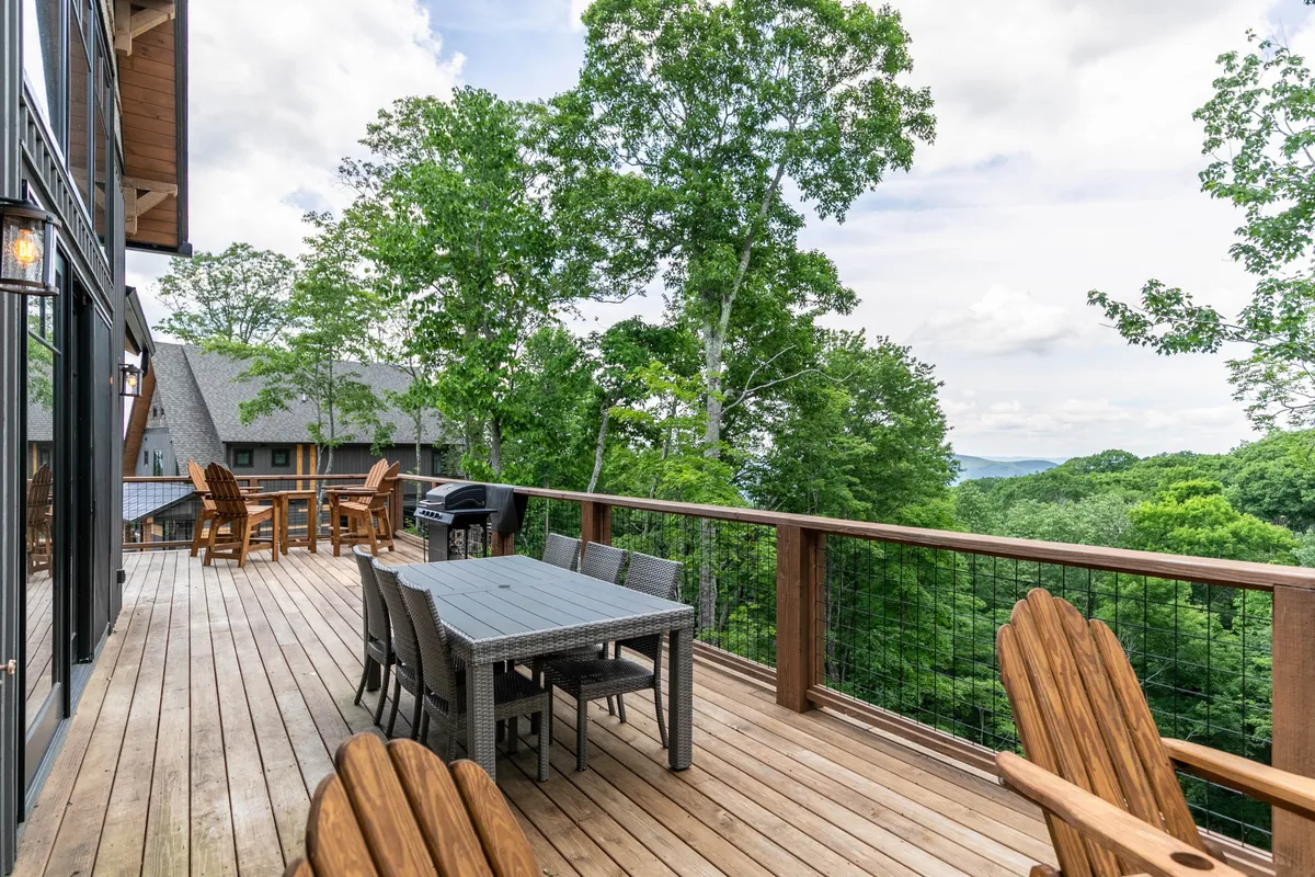 A spacious deck with a dining table and Adirondack chairs overlooks lush greenery and distant mountain views, providing an inviting setting for outdoor dining, relaxation, and family gatherings.