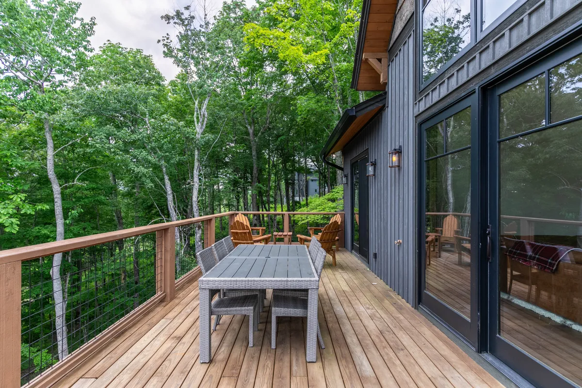 A wooden deck furnished with a dining table and Adirondack chairs, surrounded by tall trees, creates an atmosphere ideal for family meals, games, or simply relaxing outdoors in a tranquil setting.