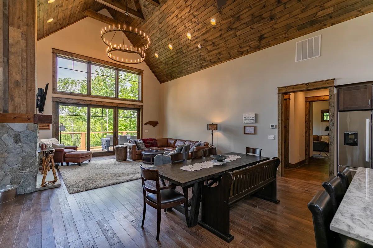 An inviting dining area that seamlessly connects with the living space, featuring a cozy seating arrangement next to large windows that frame lush greenery. The table is set for gatherings, and the ambient lighting and fireplace contribute to a warm, sociable atmosphere ideal for family meals and casual get-togethers.