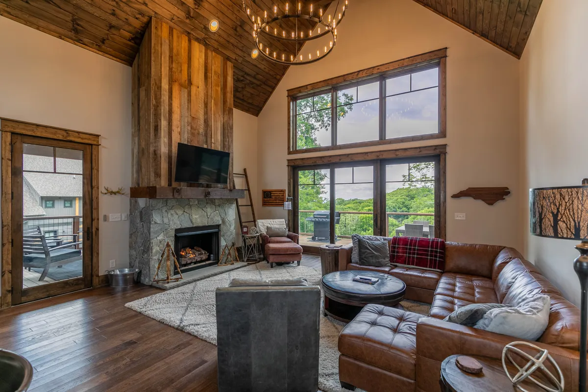 A spacious, open-concept great room featuring a striking stone fireplace as the focal point, flanked by tall windows that fill the space with natural light. The room is furnished with a plush leather sofa and highlighted by a dramatic chandelier overhead, all set against the backdrop of mountain views. The seamless connection to adjacent dining and kitchen areas is visible, emphasizing the communal and inviting atmosphere.