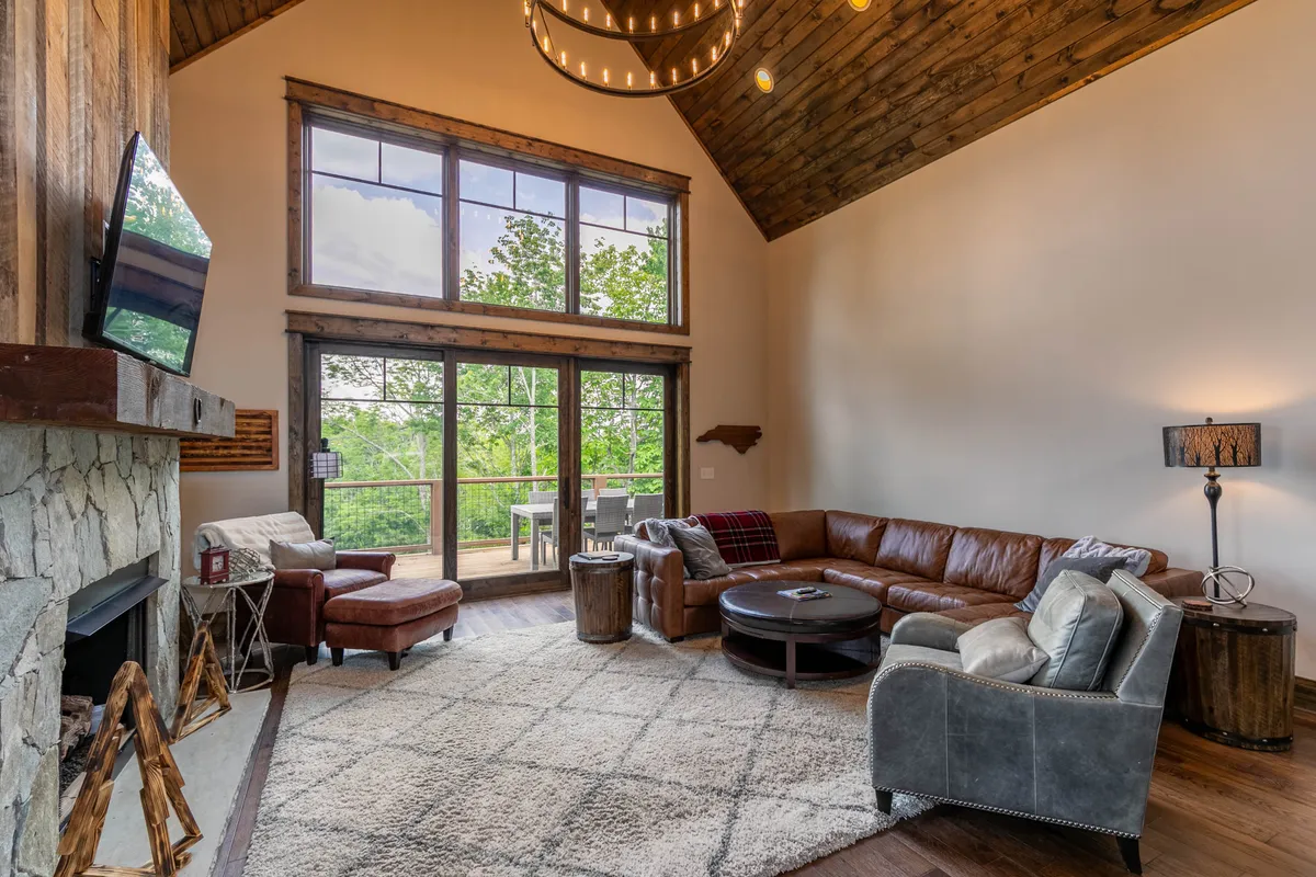 A spacious open-plan living room featuring a large leather sofa, a striking stone fireplace, and expansive windows that flood the room with natural light. The chandelier and comfortable seating create an inviting space geared toward group gatherings and shared moments.