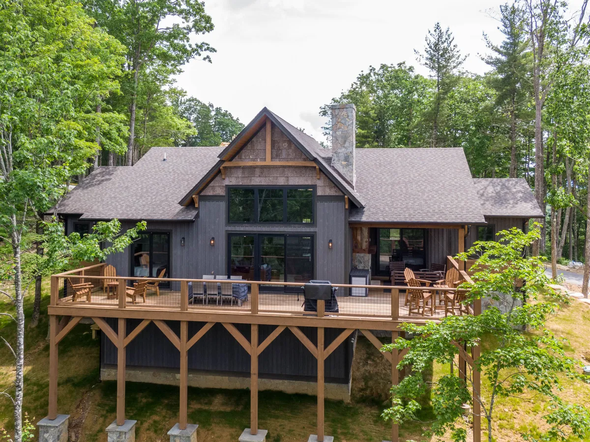 A view of the home's exterior highlights large windows, an expansive wooden deck, and a stone chimney, all nestled among trees. The deck and windows emphasize the connection between indoor comfort and outdoor splendor.